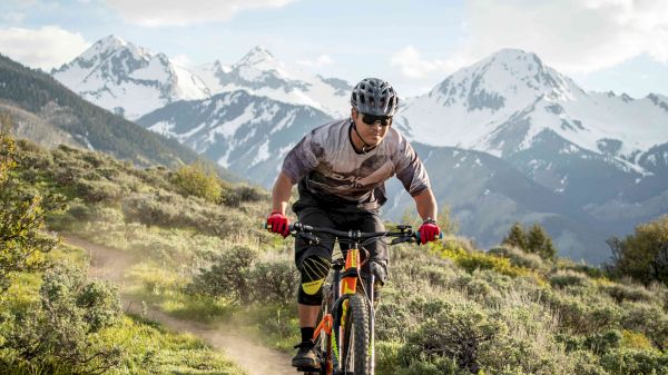 A cyclist rides a mountain bike on a trail surrounded by greenery, with snow-capped mountains in the background under a partly cloudy sky.