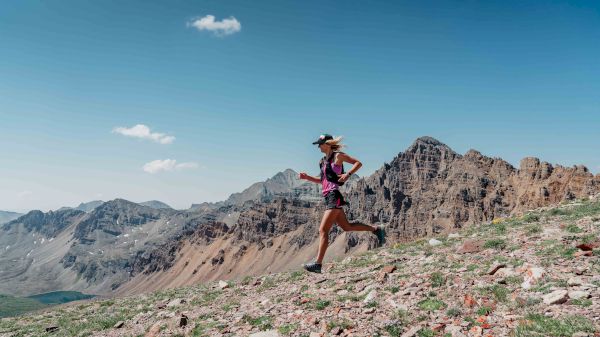 A person is trail running on a rocky mountain slope with rugged peaks and a clear sky as the backdrop.