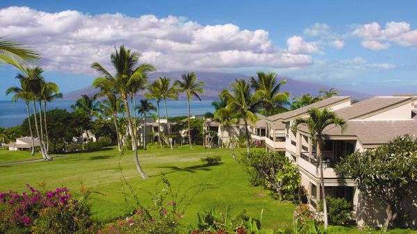 A sunny tropical resort scene with green lawns, palm trees, and beige buildings overlooking the ocean; bright sky with scattered clouds.