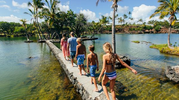 A group of people walks along a narrow stone path over clear turquoise water, surrounded by palm trees and bright blue skies.