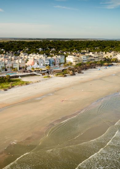 Aerial view of a sandy beach with gentle waves, lined with beachfront houses and buildings, dense greenery in the background.