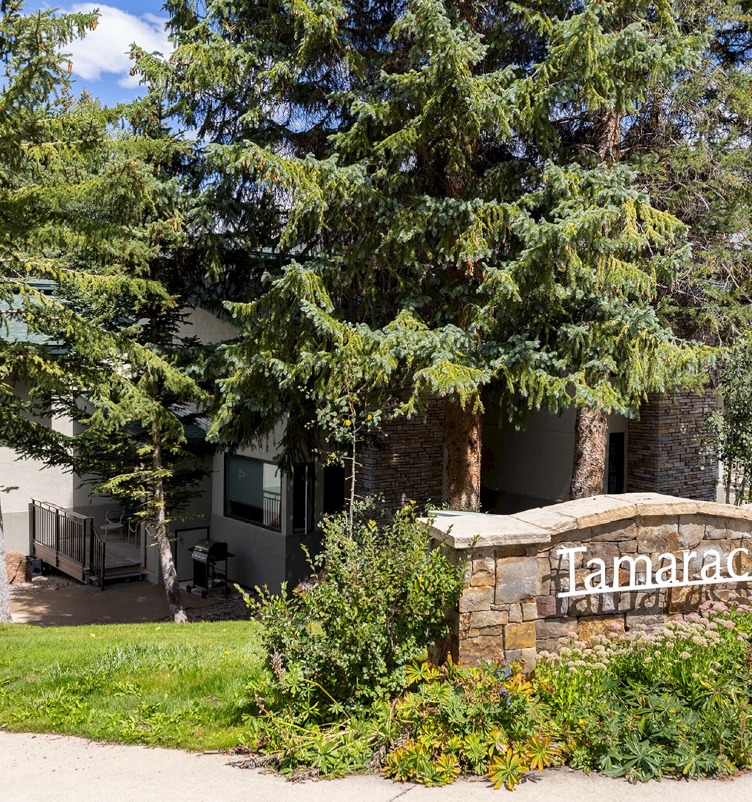 A sign reading "Tamarack" sits among trees and greenery, with a building partially visible in the background.