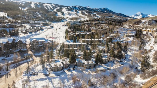 A snowy mountainous landscape with ski slopes, residential buildings, trees, and clear blue skies.