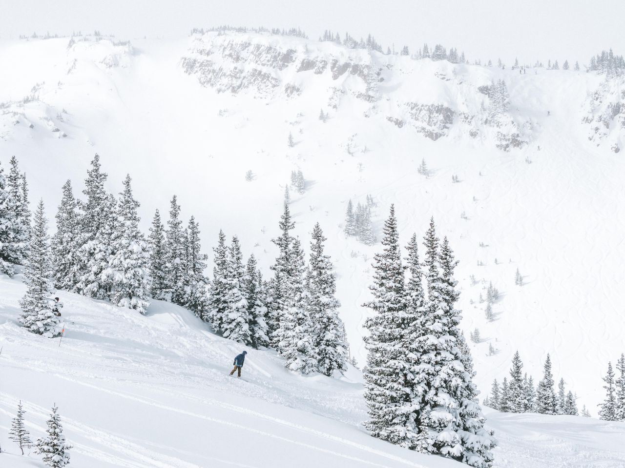 A skier navigates down a snowy slope surrounded by snow-covered trees and distant cliffs in a winter landscape.