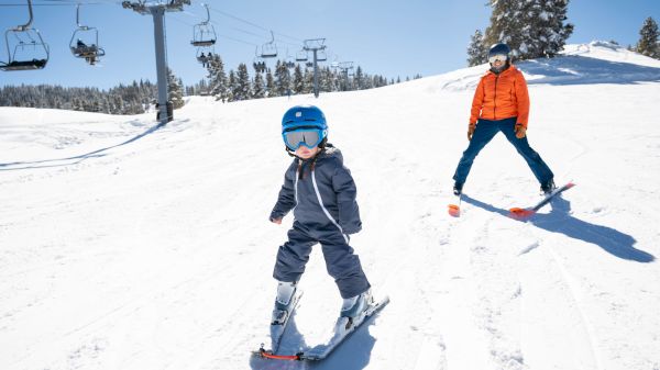 A child in ski gear is skiing down a snowy slope with an adult nearby, under a clear blue sky with ski lifts in the background.