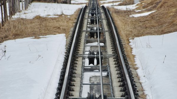 A funicular railway track is surrounded by snow, leading through a hilly landscape with trees on either side, creating a scenic winter view.
