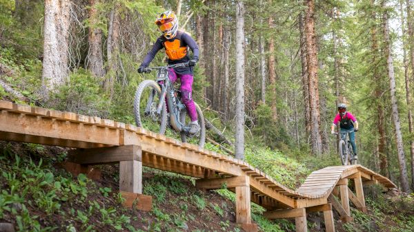 Two mountain bikers ride along a wooden trail in a forest, with trees and greenery around them.