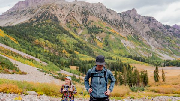 Two people hiking in a mountainous landscape with autumn colors, wearing casual outdoor clothing, under a cloudy sky.