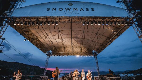 A band performs on an outdoor stage under a canopy labeled "SNOWMASS" with a scenic evening sky in the background.