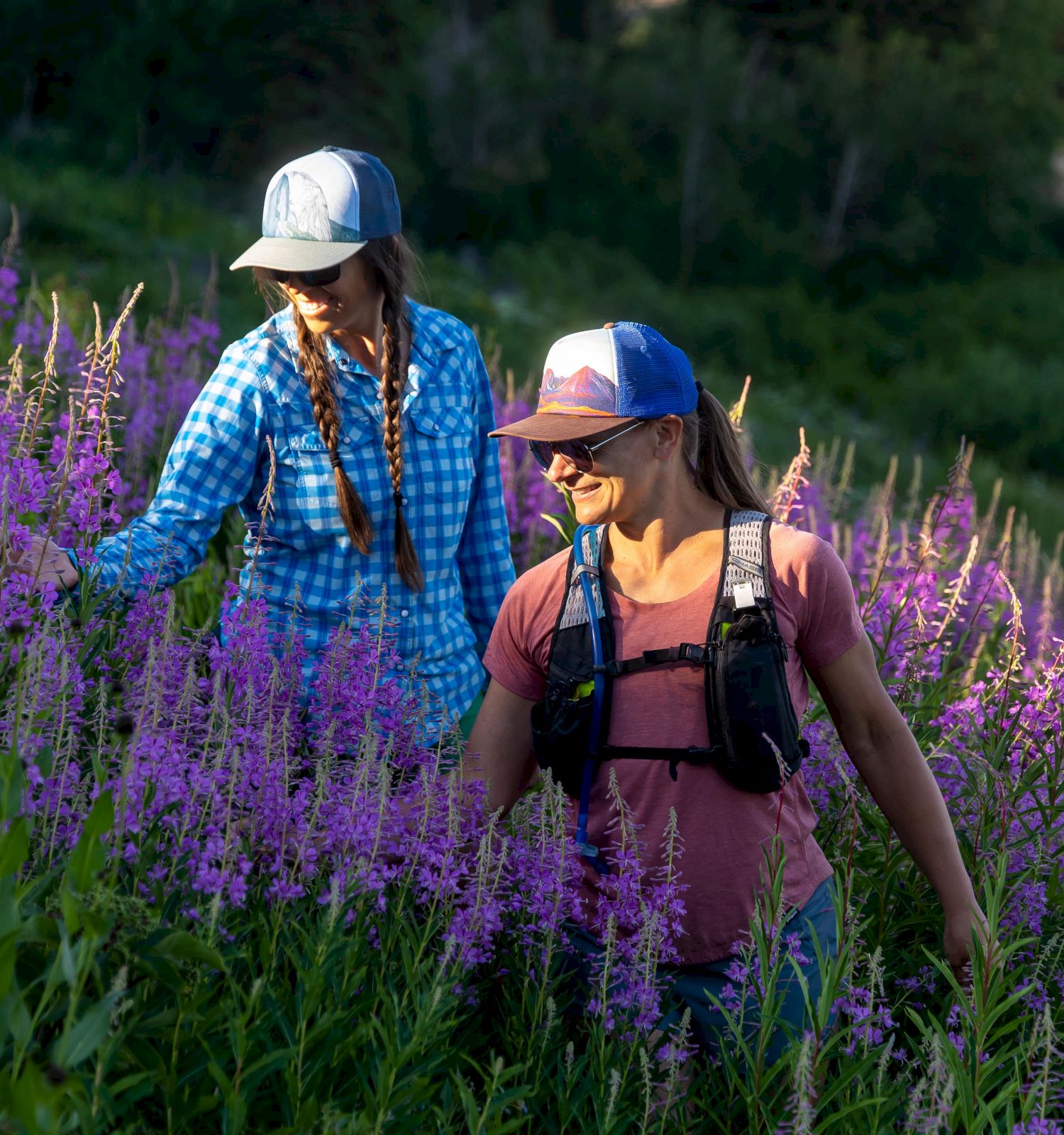 Two people in hats walk through dense purple flowers and greenery under sunlight, enjoying the outdoors.