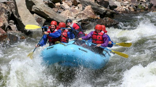 A group of people wearing helmets and life jackets is white-water rafting in a rapid river with rocky surroundings.