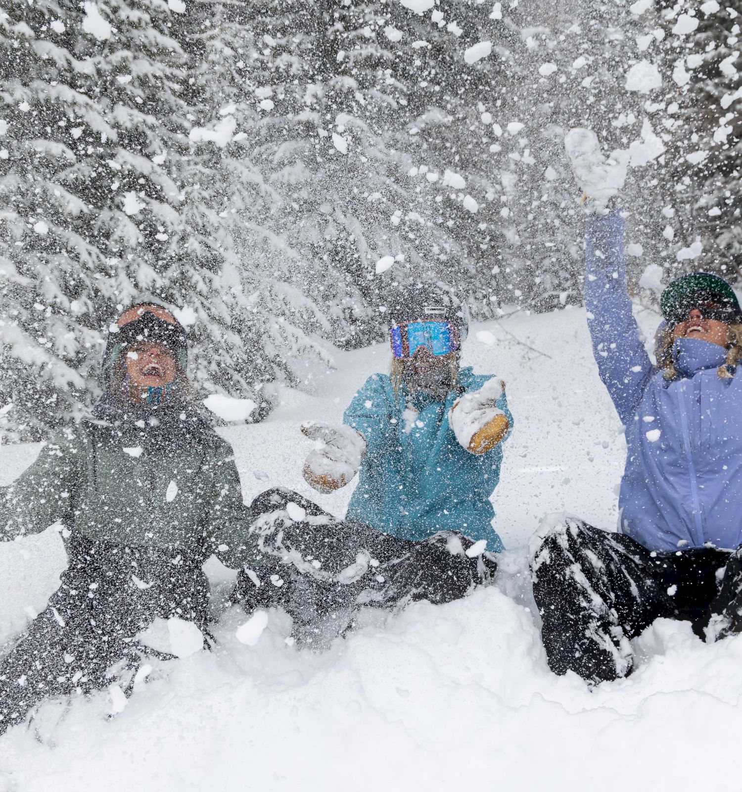 Three people sitting in the snow, playfully throwing snow in the air, surrounded by snow-covered trees.