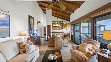 A cozy living room with sofas, chairs, a coffee table, ceiling fan, and large windows, leading to a kitchen and dining area.