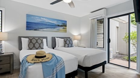 The image shows a bedroom with two single beds, a hat, and a shawl on one bed. There&rsquo;s a ceiling fan and a sliding door leading outside.