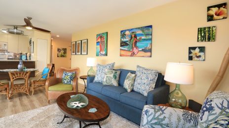 A cozy living room with a blue sofa, patterned chairs, tropical-themed decor, wall art, a wooden coffee table, and a dining area.
