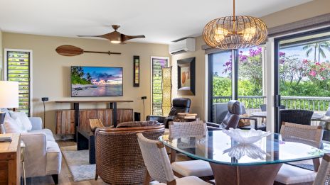 A cozy living room with wicker chairs, a sofa, a TV, art, large windows, and a glass dining table, offering a view of a garden.