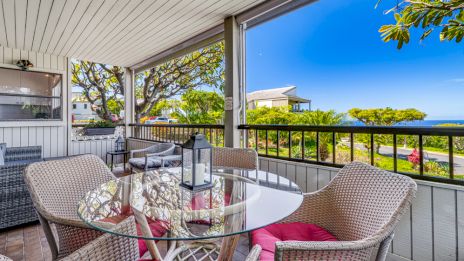 A covered patio features a glass table with red cushioned chairs, overlooking a scenic view of trees, houses, and a distant ocean.