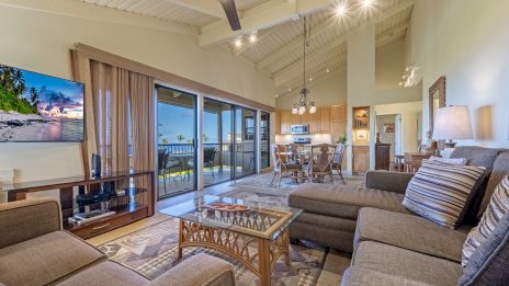 A spacious living room with a modern design, comfy seating, a flat-screen TV, and a balcony view leading to a dining and kitchen area.