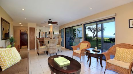 A cozy living room with wicker chairs, a sofa, and a central table, opening to a garden view through sliding glass doors.