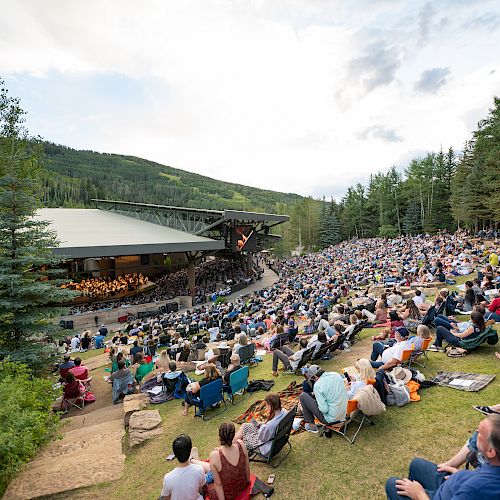 A large outdoor audience watches a concert at an open-air amphitheater surrounded by trees and mountains in daylight.
