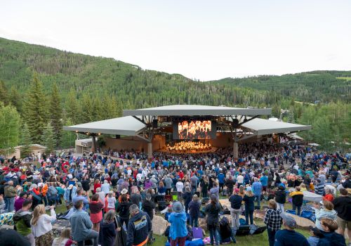 An outdoor concert amidst lush green hills, with a large audience gathered around a stage.