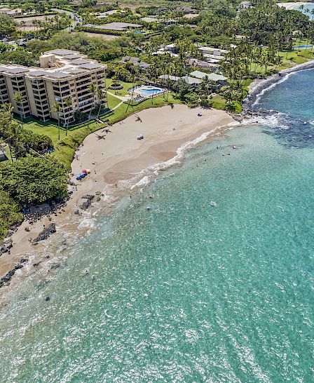 A coastal aerial view shows a beach with turquoise water, nearby buildings, and lush greenery in the surrounding area.