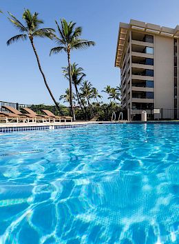 A clear blue swimming pool with lounge chairs and palm trees, in front of a multi-story hotel. The sky is partially cloudy.