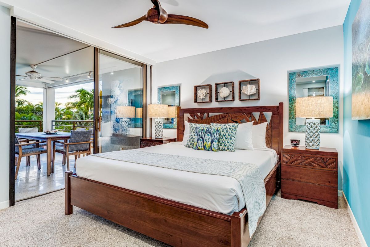 A modern bedroom with a wooden bed, decorative pillows, lamps, and art. There's access to a balcony with outdoor seating and greenery visible.