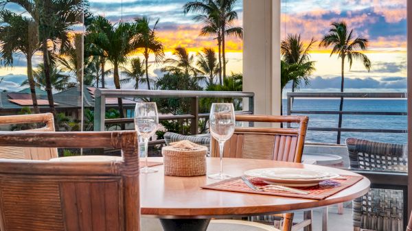 A dining table set for a meal on a balcony, overlooking a tropical sunset with palm trees and the ocean in the background.