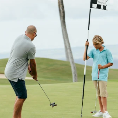 A person is putting on a golf course while a child holds the flagstick. The scene is outdoors with a distant ocean view and palm trees.