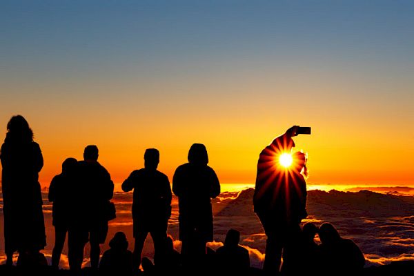 Silhouettes of people watching a sunset above clouds, with one person taking a selfie.