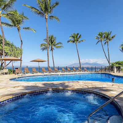 A pool area with a jacuzzi, surrounded by lounge chairs, palm trees, and a building, offers a scenic ocean view under a clear blue sky.