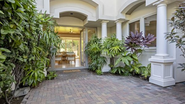 A paved entrance with plants leads to a house with white pillars and open doors, revealing a bright, inviting interior.