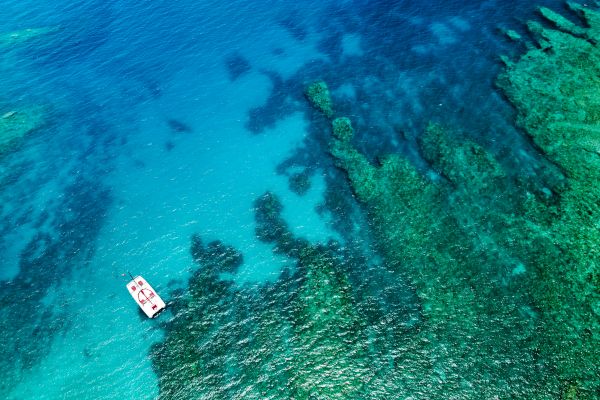 An aerial view of a boat on clear turquoise waters above a vibrant coral reef on a sunny day.