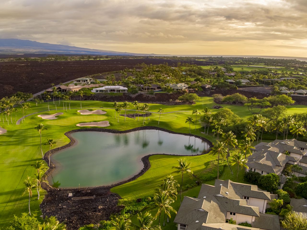 Aerial view of a lush golf course with a pond, surrounded by trees and houses, under a partly cloudy sky during sunset or sunrise.