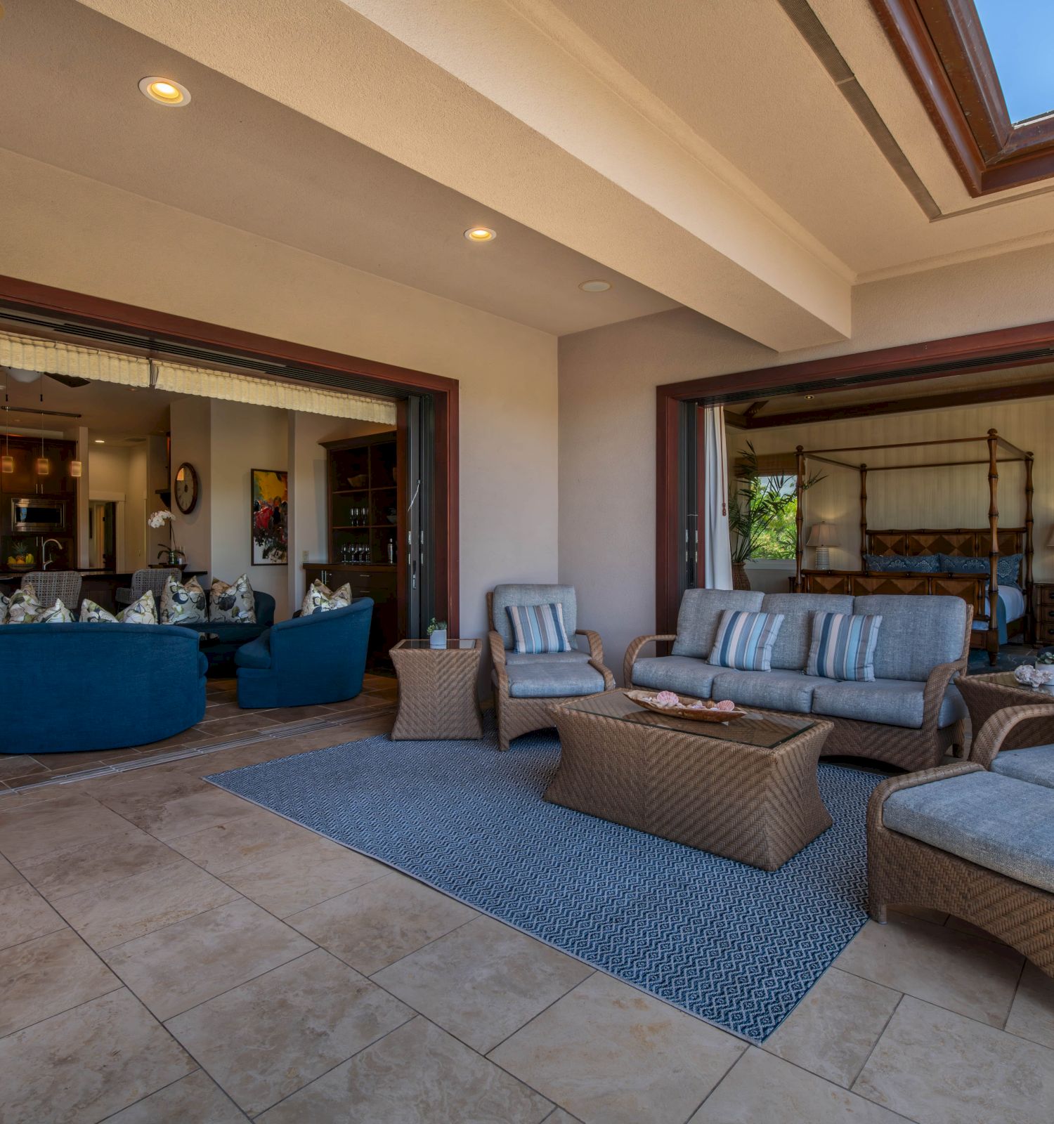 The image shows an open living space with wicker furniture, a kitchen, and a dining area. Sliding glass doors connect to an outdoor area.