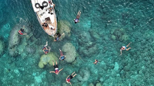 Aerial view of people snorkeling near a boat in clear turquoise water, surrounded by visible underwater rocks and coral formations.