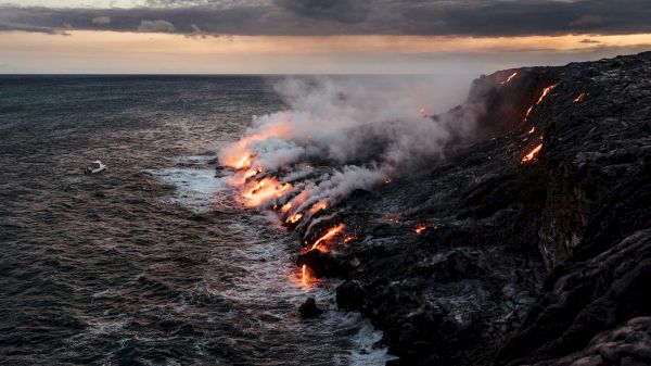 Lava flows into the ocean, creating steam and dramatic volcanic scenery with a dark, cloudy sky above.