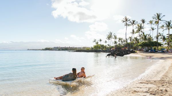 Two people relax on a lounge chair in shallow water at a sunny beach with palm trees and clear skies in the background.