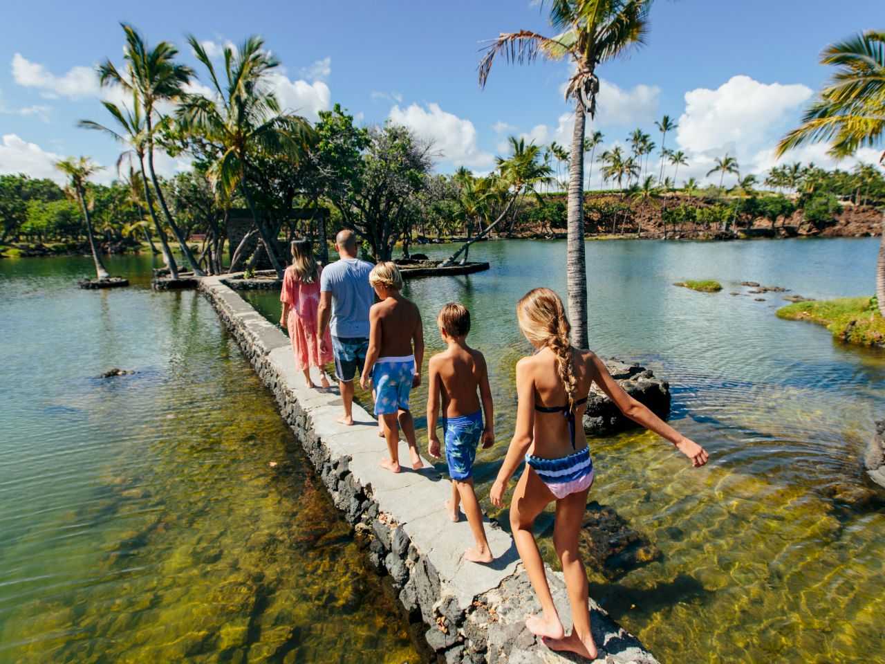 A group of people, including children, walking on a narrow stone path over water, surrounded by palm trees and sunny skies.