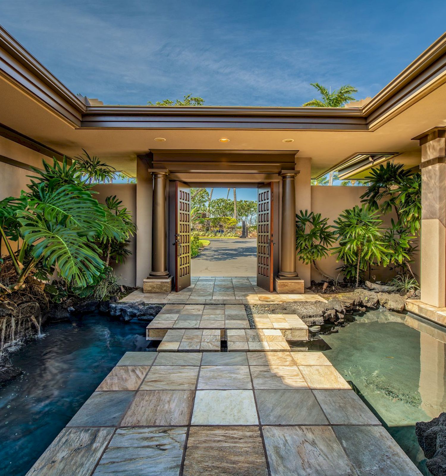 A serene entrance with a pathway over water features, surrounded by lush plants and columns, leading to a glass door and open sky.