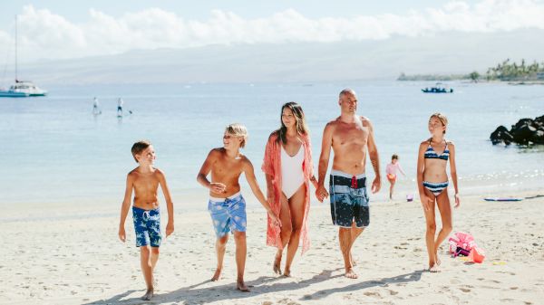 A family of five is walking on a sunny beach, wearing swimsuits, with the ocean and a boat in the background.