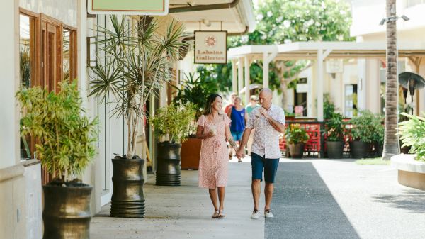 A couple walks along a shopping area, surrounded by plants and store signs, enjoying their day in a sunny outdoor setting.