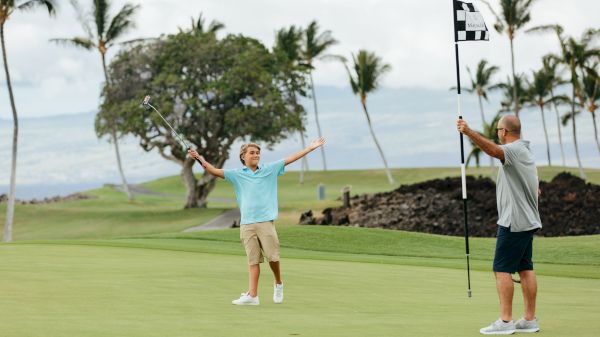 A young person celebrates on a golf course while an adult holds a checkered flag. Palm trees and a mountain are in the background.