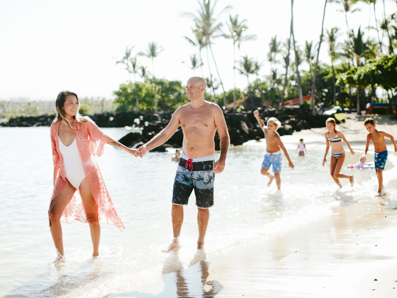 A group of people enjoy walking and playing on a sunny beach, surrounded by palm trees and clear water.