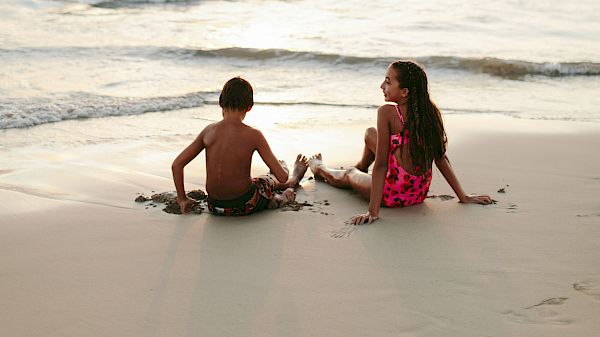 Two children sitting on a beach, with the ocean and a cloudy sky in the background.