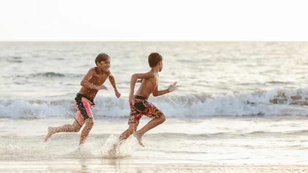 Two boys are running through the shallow waves on a beach, enjoying the sunny day and splashing water as they move.