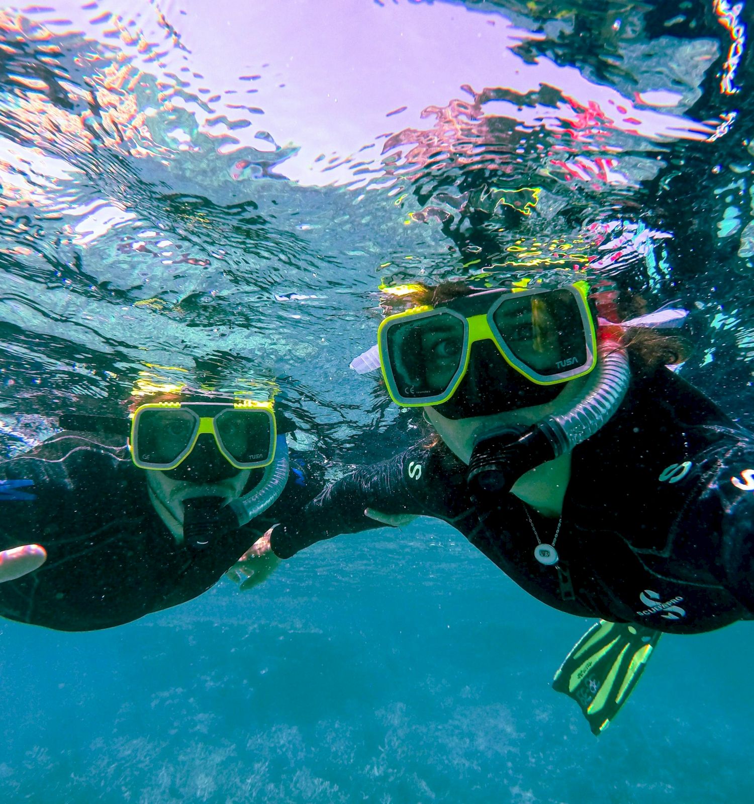 Two people snorkeling underwater, wearing yellow masks and black wetsuits, surrounded by clear blue water and reflecting sunlight.