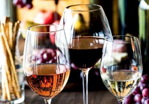 Three wine glasses with rose, red, and white wine are on a table, accompanied by breadsticks and grapes, creating a cozy ambiance.