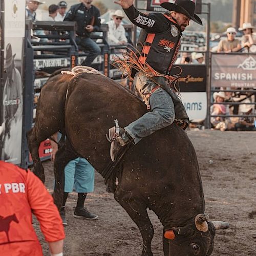 A rodeo scene with a cowboy riding a bucking bull, surrounded by onlookers and event staff, highlighting the dynamic action.